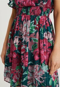 Květinové šaty se smockovaným pasem, vyrobené ze zelené látky s velkými růžovými a bílými květinami a s plisovanou strukturou.