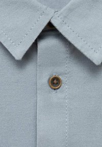 Vaaleansininen tekstiilipaita, jossa on näkyvät saumat ja yksi ruskea nappi kiinnitettynä keskelle kaulusta.