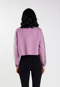 Pullover scurt, de culoare lila, cu mâneci texturate, decorat cu paiete. Are o croială lejeră și margini ribbate, asortat cu pantaloni negri ajustați.
