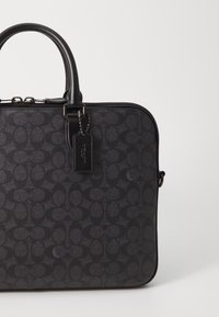 Black coated canvas bag featuring a subtle logo pattern, rectangular shape, top handles, and a branded tag attached to the zipper.