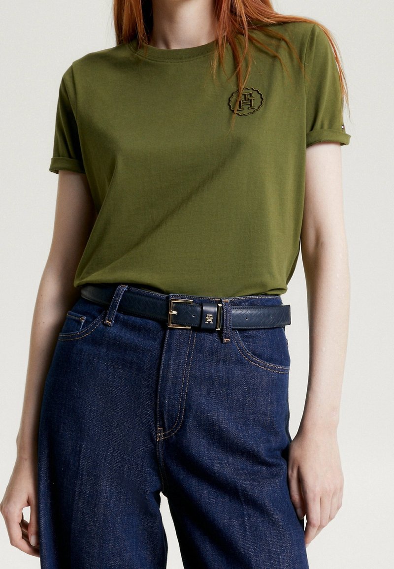 Olivgrön bomullströja med rullade ärmar och broderad logotyp. Kombinerad med mörkblå denimjeans och ett svart bälte med spänne.