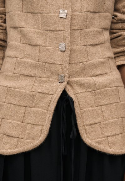 Beige gebreid vest met vierkant patroon en drie zilveren rechthoekige knopen, gedragen over zwarte broek met trekkoord in de taille.