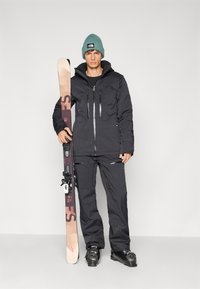 Giacca e pantaloni da sci neri isolati, con numerose tasche con zip e una finitura texturizzata, abbinati a un cappello teal e a degli sci.