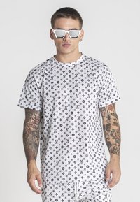Camiseta de algodón blanca con un patrón de logotipos negros repetidos. Cuello redondo, mangas cortas, corte relajado, combinada con gafas de sol blancas.