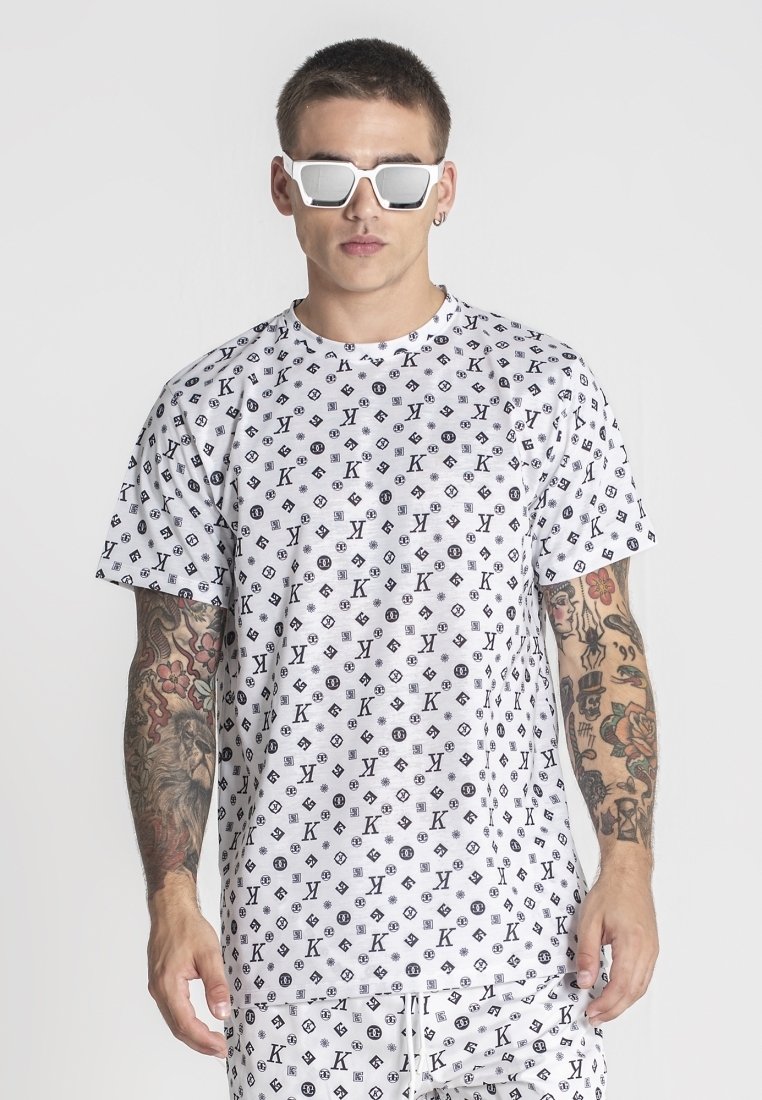 Camiseta de algodón blanca con un patrón de logotipos negros repetidos. Cuello redondo, mangas cortas, corte relajado, combinada con gafas de sol blancas.