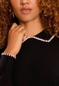 Svart stickad topp med rosa scallopkrage och manschettdetaljer. Modellens hand vilar nära halsringningen och visar den texturerade tyget.