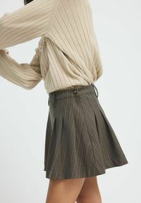 Brauner Nadelstreifenrock mit Falten und Knopfverschluss, kombiniert mit einem gerippten beigen Pullover, der die strukturierte Strickqualität und das körpernahe Design betont.
