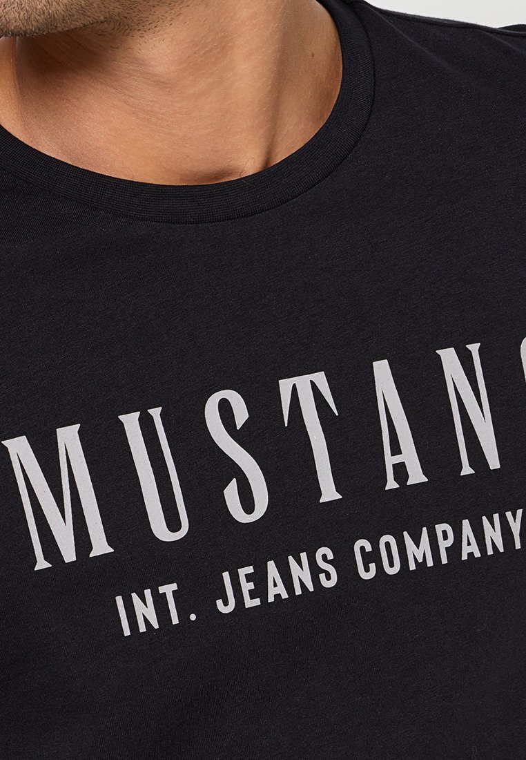 Czarna bawełniana koszulka z okrągłym dekoltem i dużym białym nadrukiem z napisem "MUSTANG INT. JEANS COMPANY" na piersi.