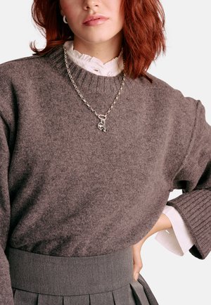 Brauner Strickpullover mit gerippten Bündchen, getragen über einer weißen Bluse. Silberne Kettenhalskette mit Anhänger. Grauer Rock mit hohem Bund.