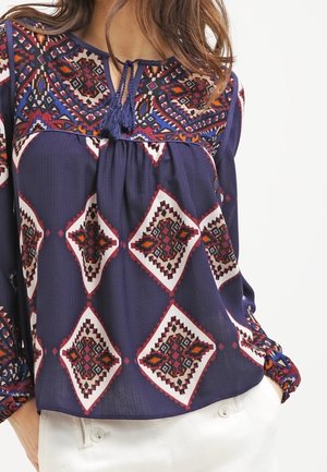 Kvinde iført en marineblå bluse med røde, hvide og orange geometriske mønstre og kvastbind ved halsudskæringen, kombineret med hvide bukser.