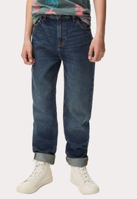 Vaqueros de mezclilla azul oscuro con un corte relajado, puños remangados y costuras en contraste; combinados con zapatillas altas blancas.