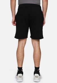 Pantalones cortos de algodón negros con una cintura elástica y bolsillos laterales. Se llevan con zapatillas blancas y calcetines negros, mostrando un ajuste informal.