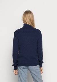 Pull en tricot côtelé marine avec un col montant, manches longues et coupe décontractée, associé à un jean bleu clair. Texture lisse, design minimaliste.