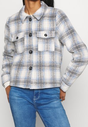 Femme portant une veste à carreaux boutonnée aux couleurs bleu, gris et blanc avec deux poches poitrine, assortie à un jean bleu.