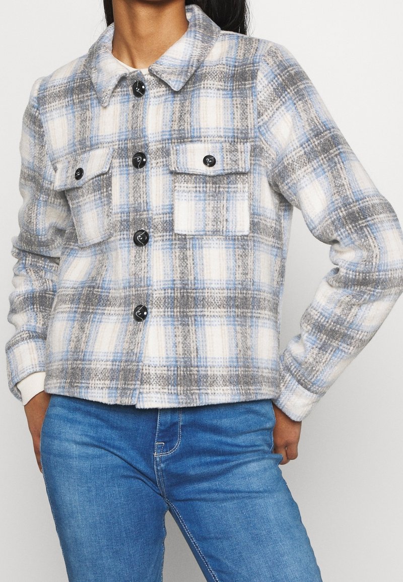 Femme portant une veste à carreaux boutonnée aux couleurs bleu, gris et blanc avec deux poches poitrine, assortie à un jean bleu.