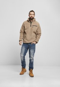 Tan fleece pullover med høj krave, lynlåslomme på brystet og sidelommer, parret med blå jeans og brune støvler.