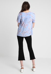Blusa floral de color azul claro con mangas cortas, que presenta un pliegue en la parte posterior y una tela suave. Combinada con unos jeans negros acampanados y sandalias blancas de tacón.