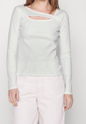 Kobieta ubrana w długi rękaw biały prążkowany top z poziomym wycięciem poniżej linii dekoltu oraz jasne spodnie.