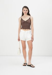 Brun sydd tilpasset tanktop med rynket tekstur og kantede kanter; paret med hvite linshorts, svarte flip-flops og en liten hvit veske.