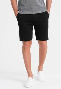 Schwarze Shorts aus glattem Stoff, ausgestattet mit seitlichen Taschen und einem flachen Vorderdesign. Kombiniert mit weißen Slippern und einem grauen T-Shirt.