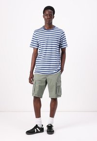 Gestreepte blauwe en witte t-shirt, groene cargoshorts, zwarte sneakers met witte accenten. Model staat tegen een effen achtergrond.