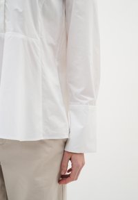 White button-up shirt with a structured design, featuring a peplum silhouette, long sleeves, and smooth, lightweight fabric.