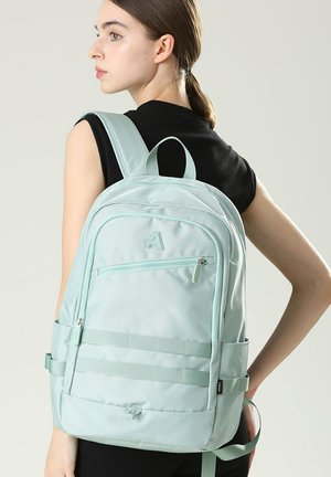 Jonge vrouw met bruin haar in een paardenstaart draagt een lichtblauwe rugzak over een zwart mouwloos shirt, kijkend naar links tegen een effen achtergrond.