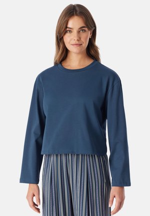 Frau mit braunen Haaren, die ein schlichtes marineblaues Langarmshirt und vertikal gestreifte Hosen trägt, steht vor einem weißen Hintergrund.