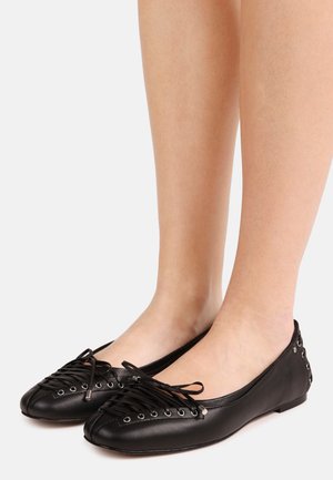 Bailarinas de cuero negro con cordones en la parte delantera y ojetes plateados, usadas en pies desnudos sobre un fondo blanco.