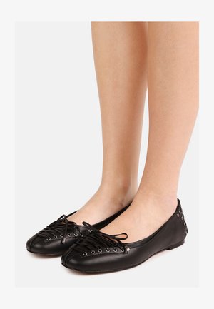 Fekete bőr balettcipő fodros fűzővel és ezüst szemekkel, mezítláb viselve, fehér háttér előtt.