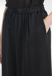 Mustat leveälahkeiset housut, joissa on kuminauhalla kerätty vyötärö, säädettävä kiristysnyöri ja sivutaskut. Housut on valmistettu sileästä, kevyestä kankaasta.