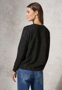 Schwarzer Sweatshirt mit geometrischem Texturmuster in einem leicht glänzenden Finish; verfügt über einen Rundhalsausschnitt und lange Ärmel, kombiniert mit blauen Jeans.