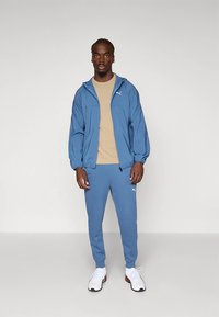 Blue lightweight jacket with hood, beige t-shirt, and matching blue tracksuit pants. White sneakers with red accents.