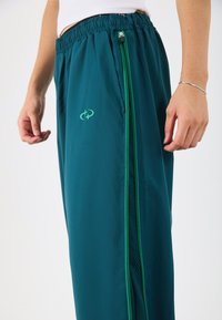 Türkise Sporthose mit elastischem Bund, grünen seitlichen Reißverschlüssen und Mesh-Einsätzen. Verfügt über ein grünes Logo auf der linken Vorderseite.