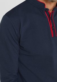 Mörkblå långärmad polo med en faux krage, som har röda detaljer och knapplist. Strukturerat tyg med en slät yta.