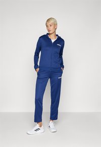 Zilā krāsā zip-up treniņtērps ar kapuci, uz krūšu ir izšūta logo. Gluda auduma, brīvi pieguļošs, saskaņots ar baltām sporta apaviem.