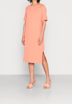 Femme portant une robe T-shirt corail longueur genou avec fente latérale et sandales à talons hauts assorties.