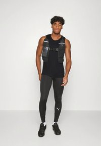 Černé atletické tílko v kombinaci s černými přiléhavými legínami a černými sportovními botami. Jedinec nosí černý postroj s více kapsami.