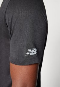 Juodas sportinis marškinėlis su tekstūruotu raštu. Ant rankovės yra pilkas New Balance logotipas. Trumpų rankovių dizainas pritaikytas komfortui.