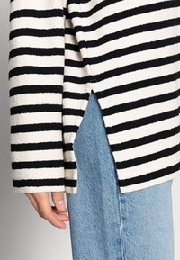 Randig stickad tröja i kräm och svart med en sidsspricka, matchad med ljusblå denim. Tyget verkar mjukt och strukturerat.