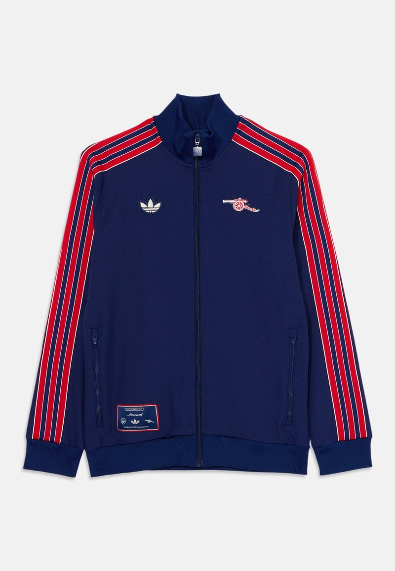 Mörkblå jacka med röda ränder, dragkedjestängning, hög krage, Adidas-logotyp på bröstet och Arsenal-kanonemblem. Har en sidoficka med dragkedja.