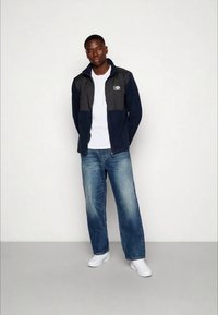 Casaco de fleece azul-marinho e cinza com zipper, t-shirt branca e jeans azuis. Modelo a usar tênis brancos, de pé com os braços relaxados.