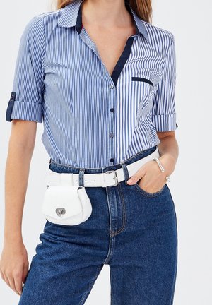 Kobieta ubrana w niebiesko-białą koszulę w pionowe paski z podwiniętymi rękawami, niebieskie dżinsy z wysokim stanem, biały pasek i biała torebka na biodra.