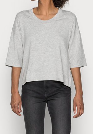 Femme portant un haut ample gris clair à manches courtes avec un jean noir moulant, vue du cou jusqu'aux hanches, sur un fond uni.