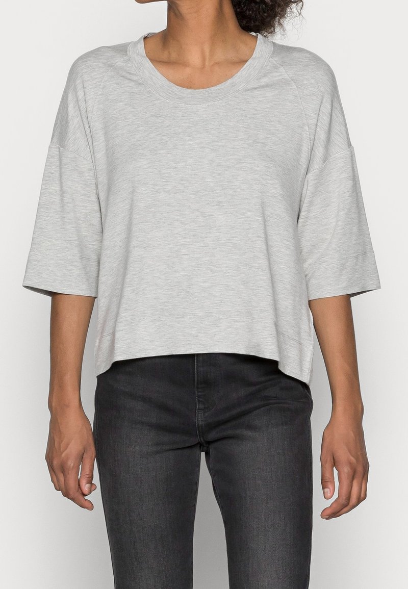 Femme portant un haut ample gris clair à manches courtes avec un jean noir moulant, vue du cou jusqu'aux hanches, sur un fond uni.