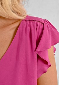 Blusa fucsia con maniche corte a volant e scollatura a V. Tessuto morbido e testurizzato con dettagli arricciati sulle spalle.