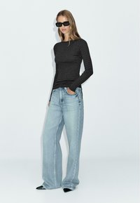 Zwarte en grijze gestreepte top met lange mouwen en een aansluitende pasvorm, gecombineerd met lichtblauwe wijde jeans. Het model draagt zwarte hoekige zonnebril en puntige hakken.