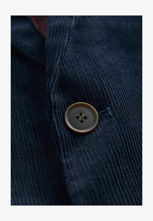 Jūras zils korduroja audums ar lielu, apaļu, brūnu pogu ar četrām caurumiem, piešūtu uz nedaudz teksturētas virsmas.