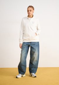 Lys beige fleece quarter-zip genser med kenguru-lomme; sammen med løstsittende middels blå denimjeans og grå joggesko.