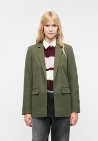 Olivovo zelený blejzer s klopou a dvoma prednými vreckami, prehnaný cez pruhovaný sveter v odtieňoch bordovej, bielej a béžovej.